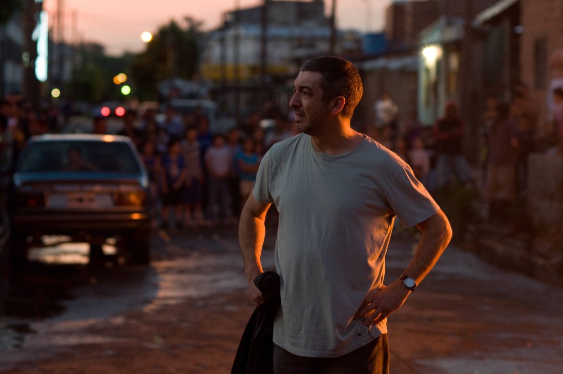 Man in light-colored shirt standing on urban street at dusk with cars and crowds in background.