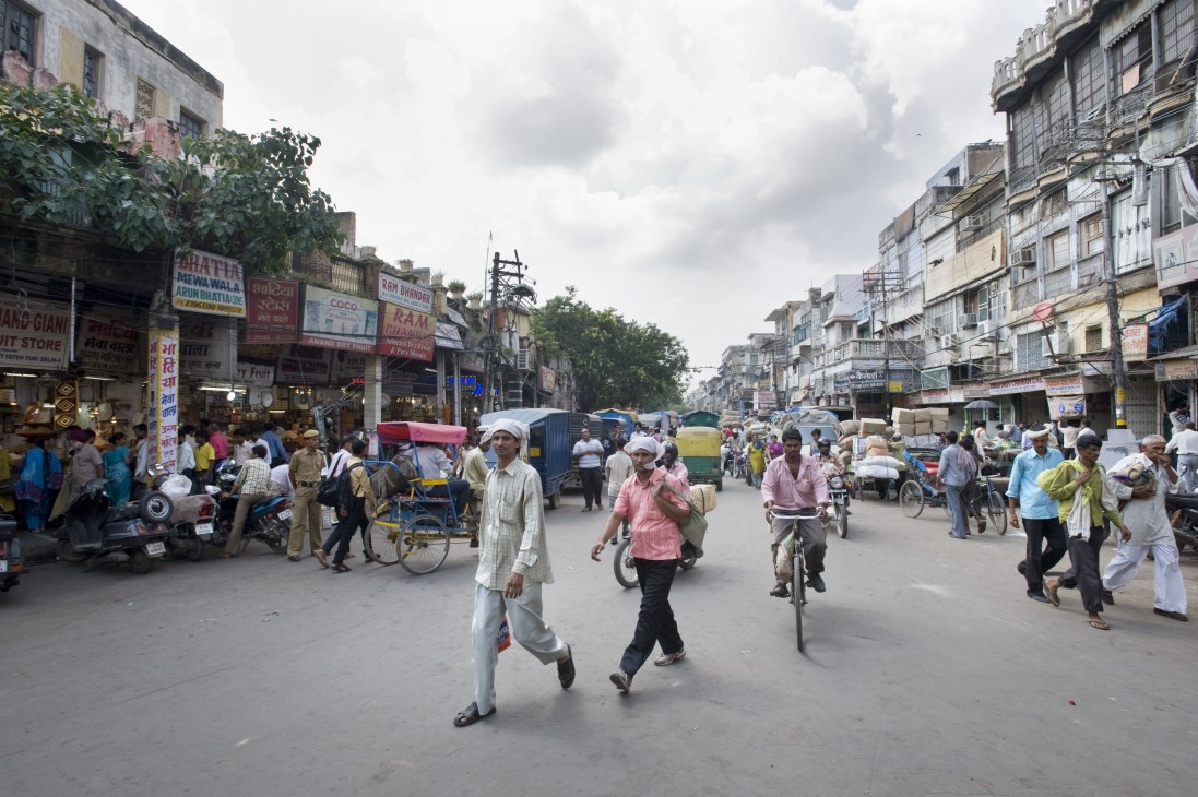 Busy Khari Baoli market street in Old Delhi with pedestrians, vendors, bicycles and colorful shop signs lining historic buildings.