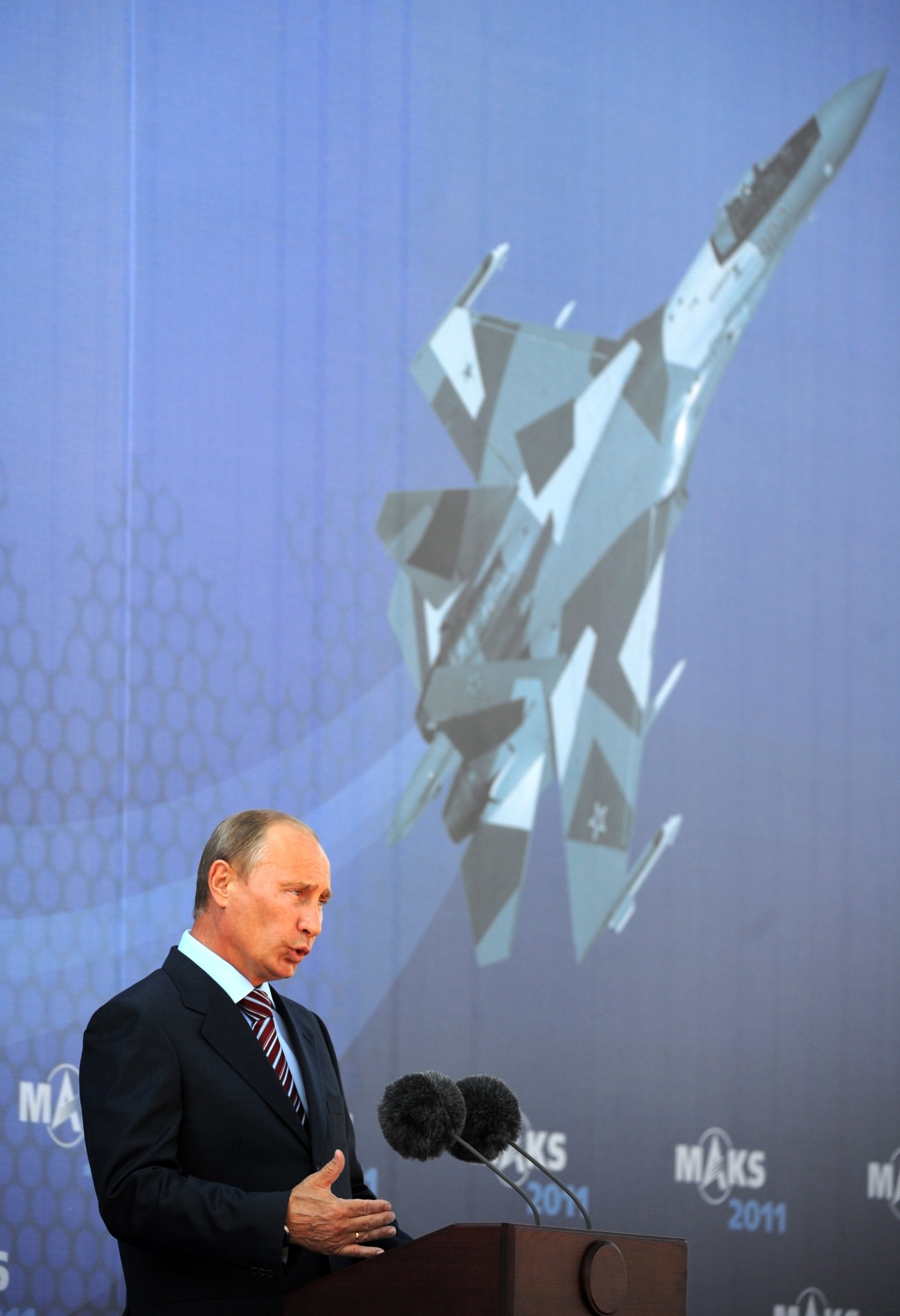 Vladimir Putin speaks at a podium with a T-50 stealth fighter jet displayed on the screen behind him.
