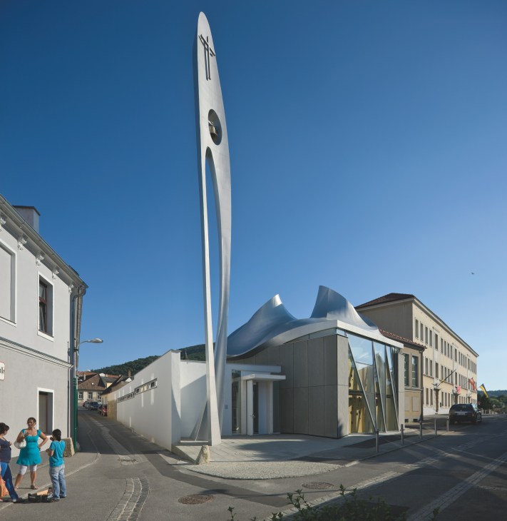Martin Luther Church in Hainburg, Austria, designed by Coop Himmelb(l)au, featuring a dramatic twisted steel bell tower rising above surrounding town buildings under clear blue sky.