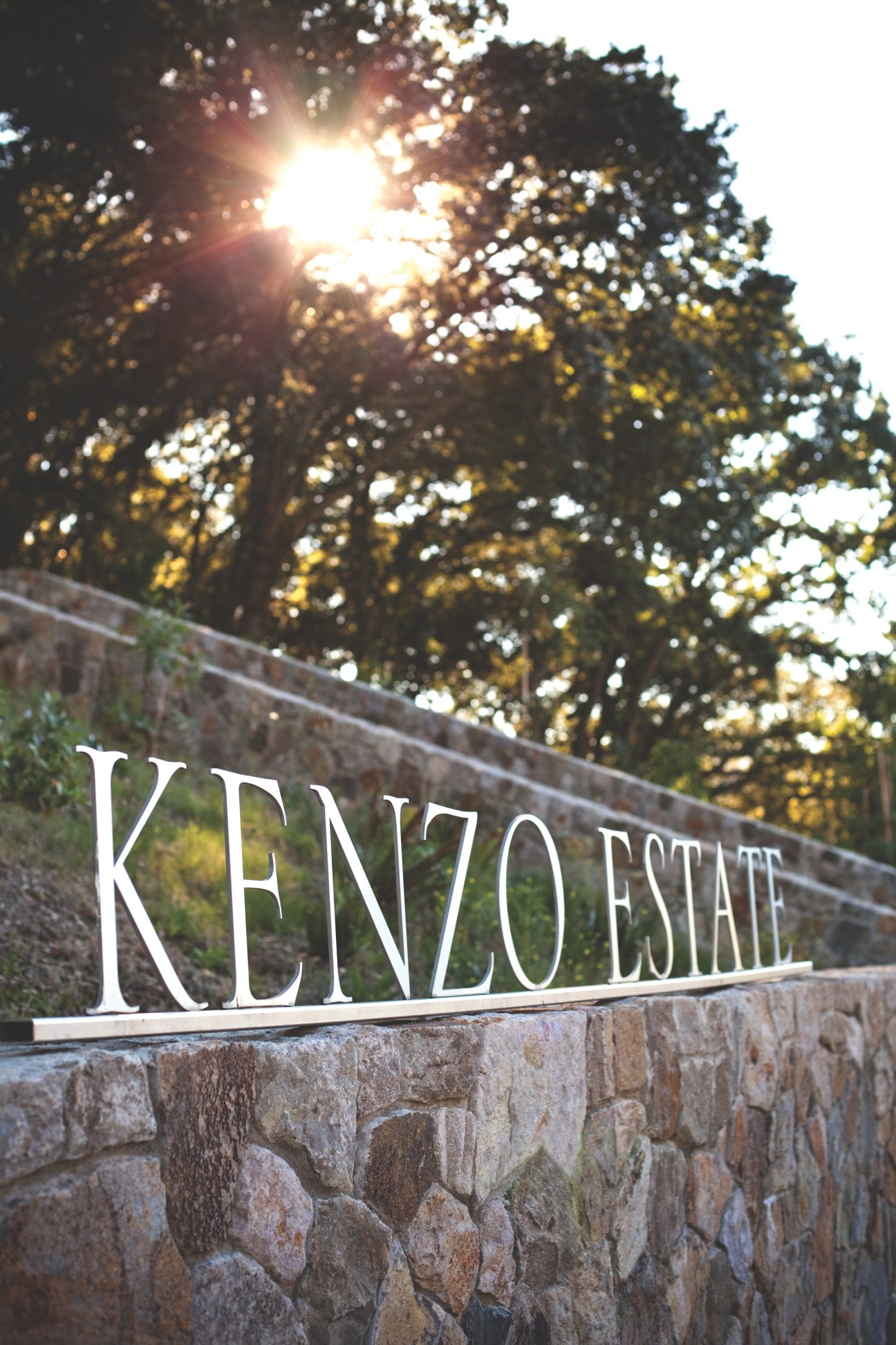 Kenzo Estate entrance sign on stone wall with trees and sun behind