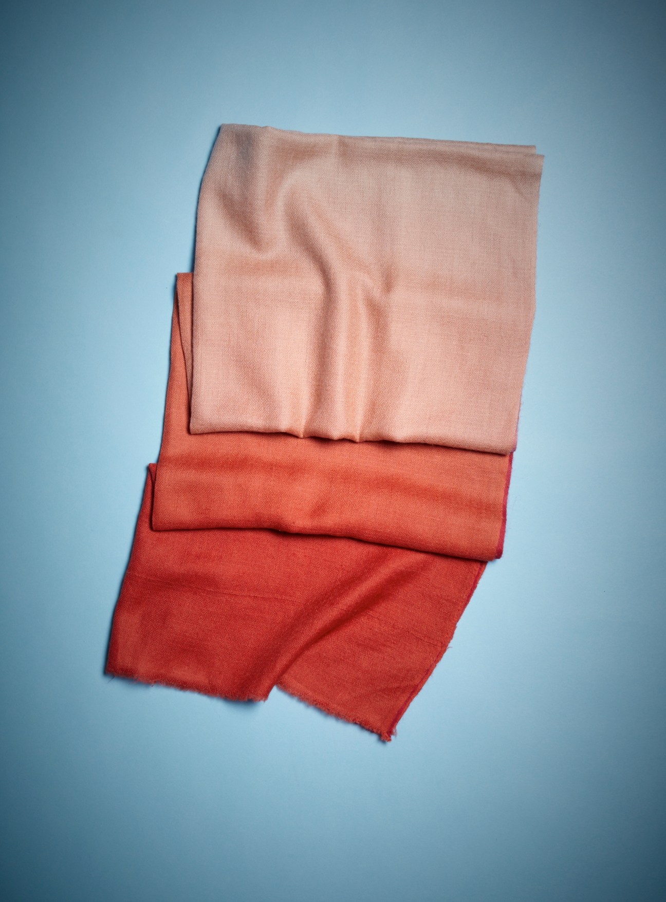 Three folded Ahilya scarves in pink, coral and red shades against a blue background.