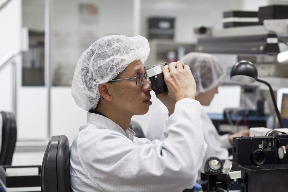 Worker in white protective gear and hairnet inspects camera equipment through a lens in a Leica manufacturing facility.
