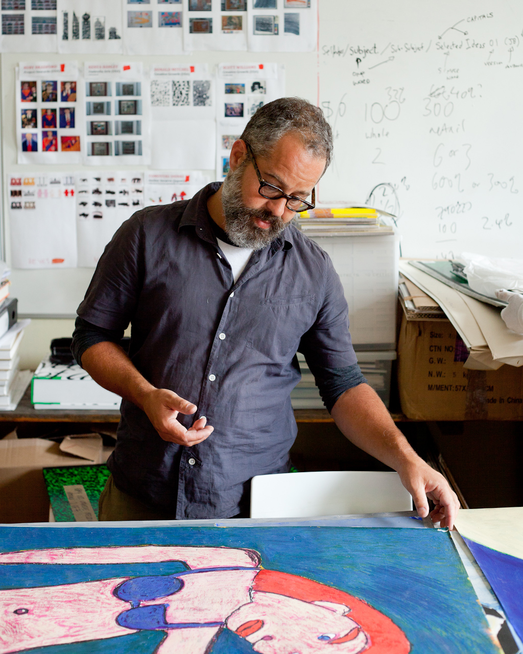 Man with glasses examining artwork at a desk surrounded by sketches and creative materials.
