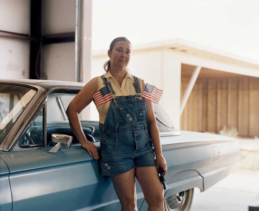 Babbi Girl, a Tea Party member, poses next to a vintage car wearing denim overalls and holding American flags.