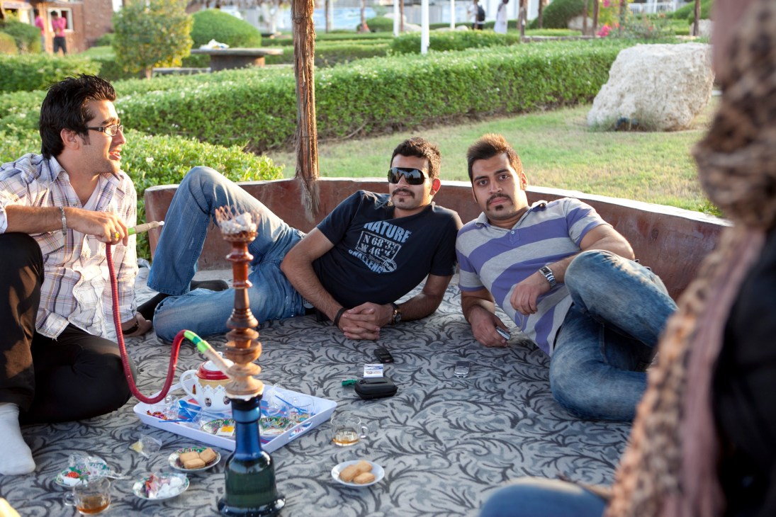 Four men sit on a bench in a park setting with food and a hookah pipe, relaxing in a landscaped garden area.