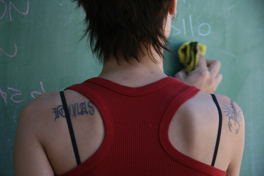 Woman with tattoos on her shoulders writing on a green chalkboard while holding chalk.