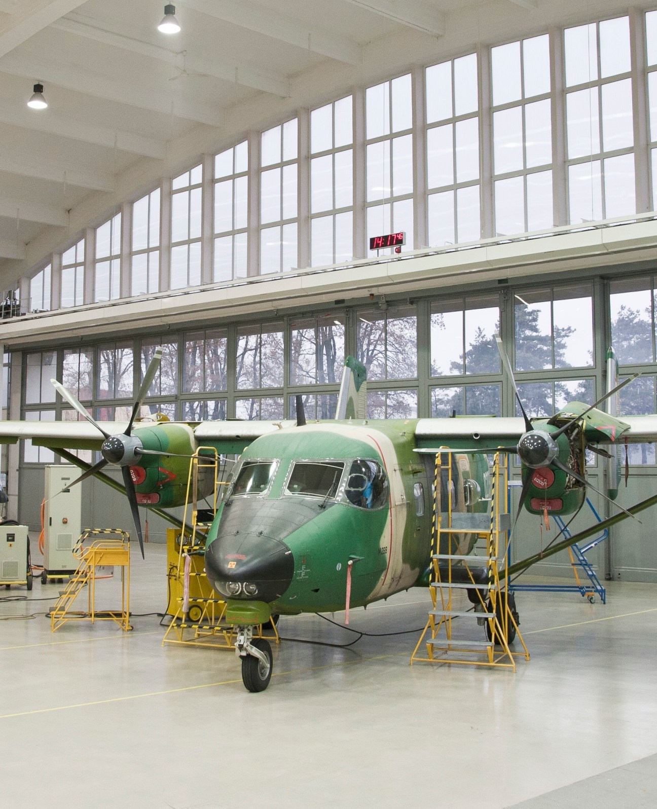 Green and grey M28 aircraft under maintenance in a bright factory hangar with large windows.