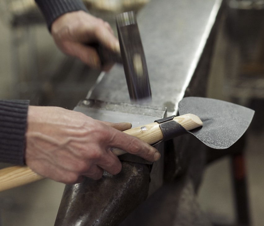 Hands attaching a wooden handle to a metal garden tool head during manufacturing.