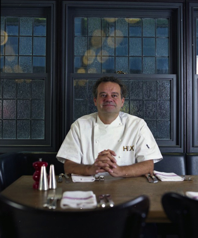 Mark Hix, restaurateur, seated at a table in his restaurant wearing a white chef's shirt with HIX branding.