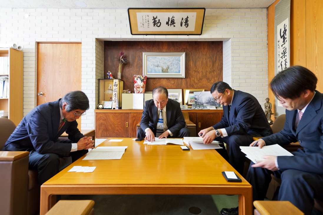 Five men in business suits review documents at a wooden table in a Japanese office with calligraphy art.