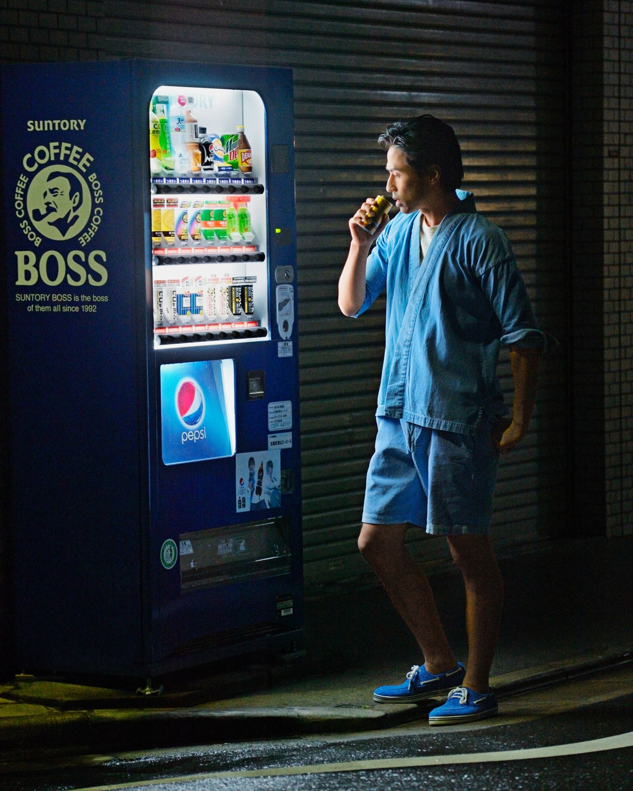 Man in denim jacket and shorts drinking from a Suntory Boss vending machine at night in Tokyo.