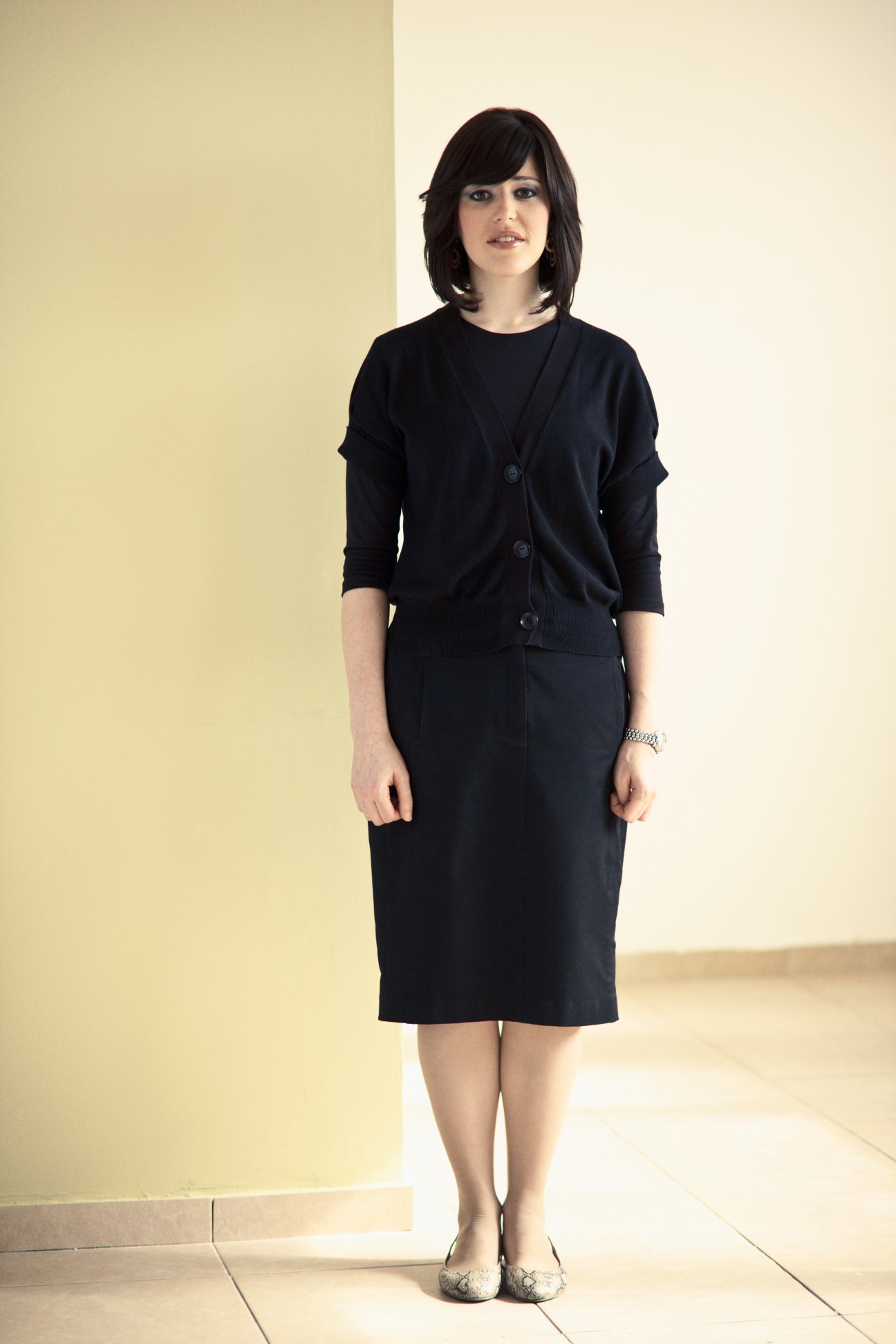 Student Debbie Englard wearing a black cardigan and dress against a pale yellow wall.
