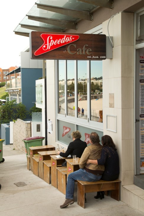 Three people seated on wooden benches outside Speedos Café at Bondi Beach, with the café's neon sign visible above.