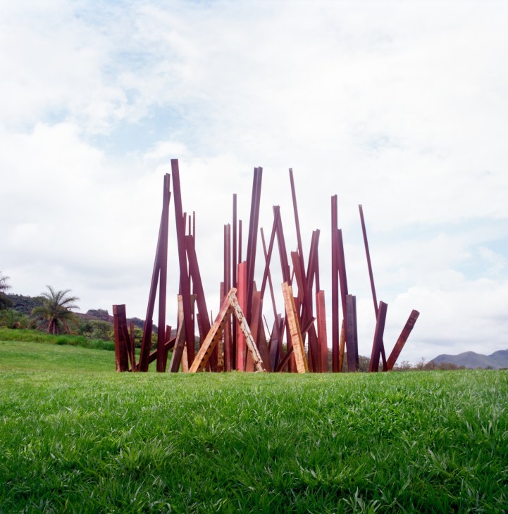 Large-scale sculptural installation by American artist Chris Burden at Inhotim art centre featuring red metal beams arranged in a dramatic angular formation in a green field.