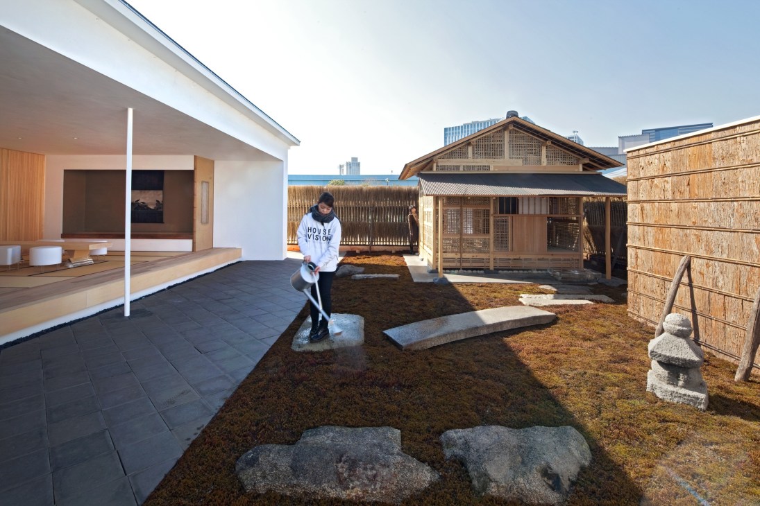 Person in white hoodie stands in courtyard between modern and traditional Japanese wooden structures with moss garden and stone features.