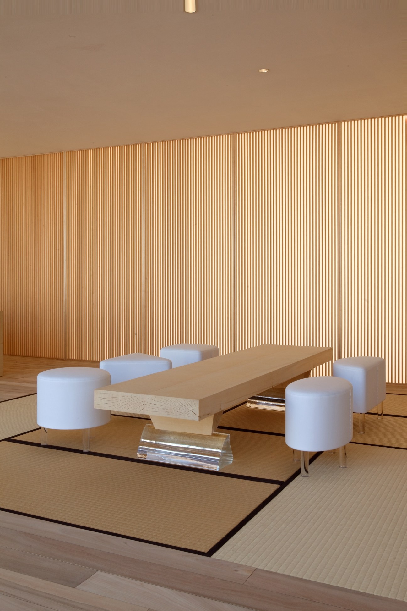 Minimalist interior with white cylindrical stools arranged around a wooden table on tatami mat flooring, with vertical wood slat wall paneling and soft warm lighting.