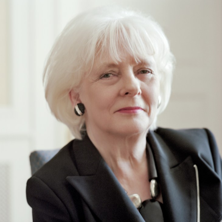 Woman with white hair wearing black trouser suit and earrings in formal portrait setting.