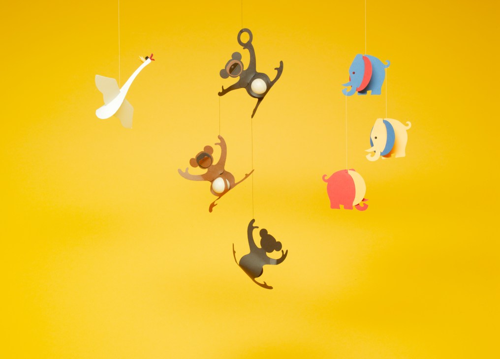 Handcrafted paper mobiles hanging against a yellow background, including monkey, swan and elephant designs by Livingly.