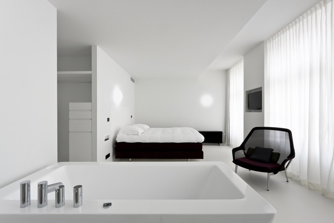 Minimalist hotel bedroom with white Corian bathtub in foreground, black bed, modern chair, and wall-mounted television at Hotel Zenden.