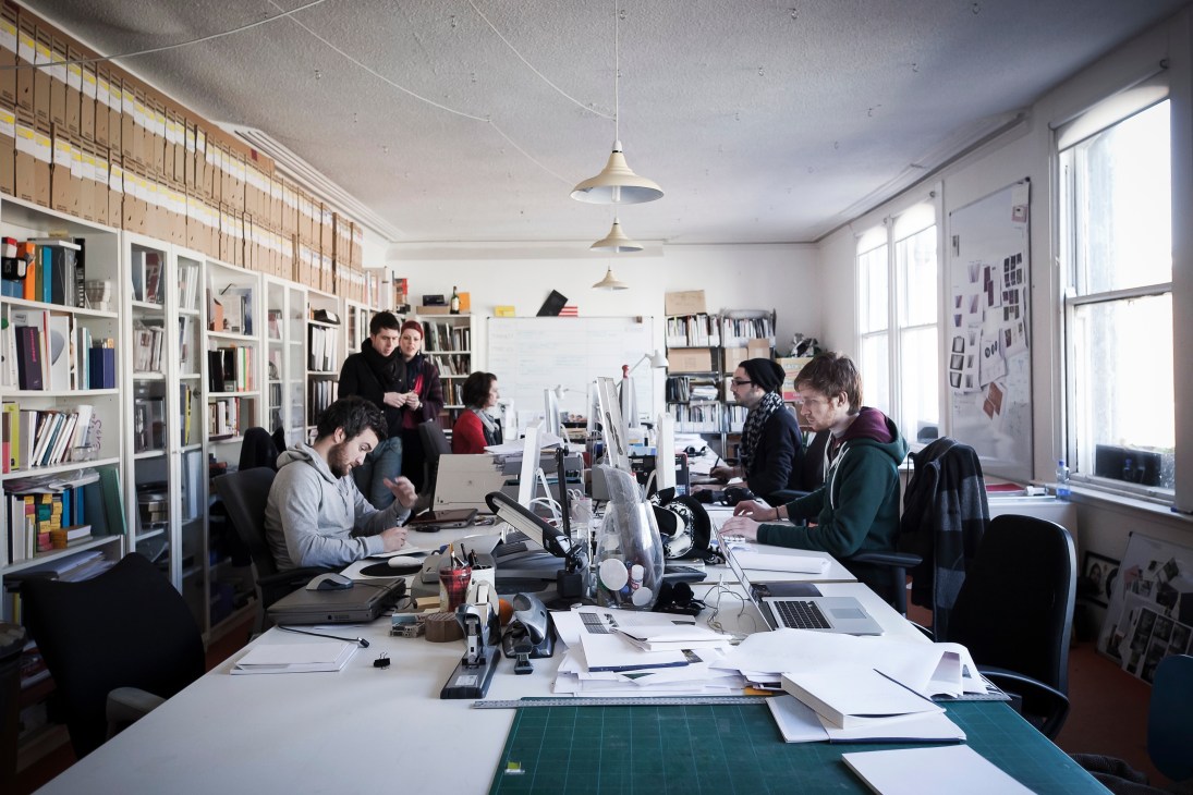 Interior of a bright design studio with multiple employees working at desks with computers, shelves of books and materials lining the walls.