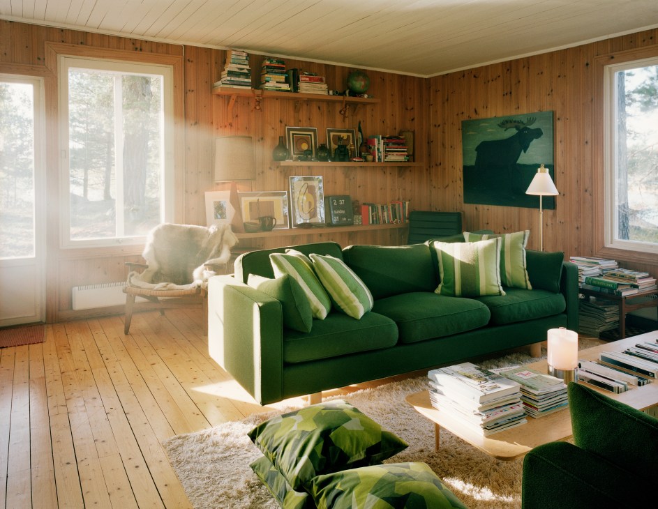 Interior of a 1960s wooden cabin with a green sofa, shaggy rug, cushions, shelves with books and art, and windows overlooking trees.