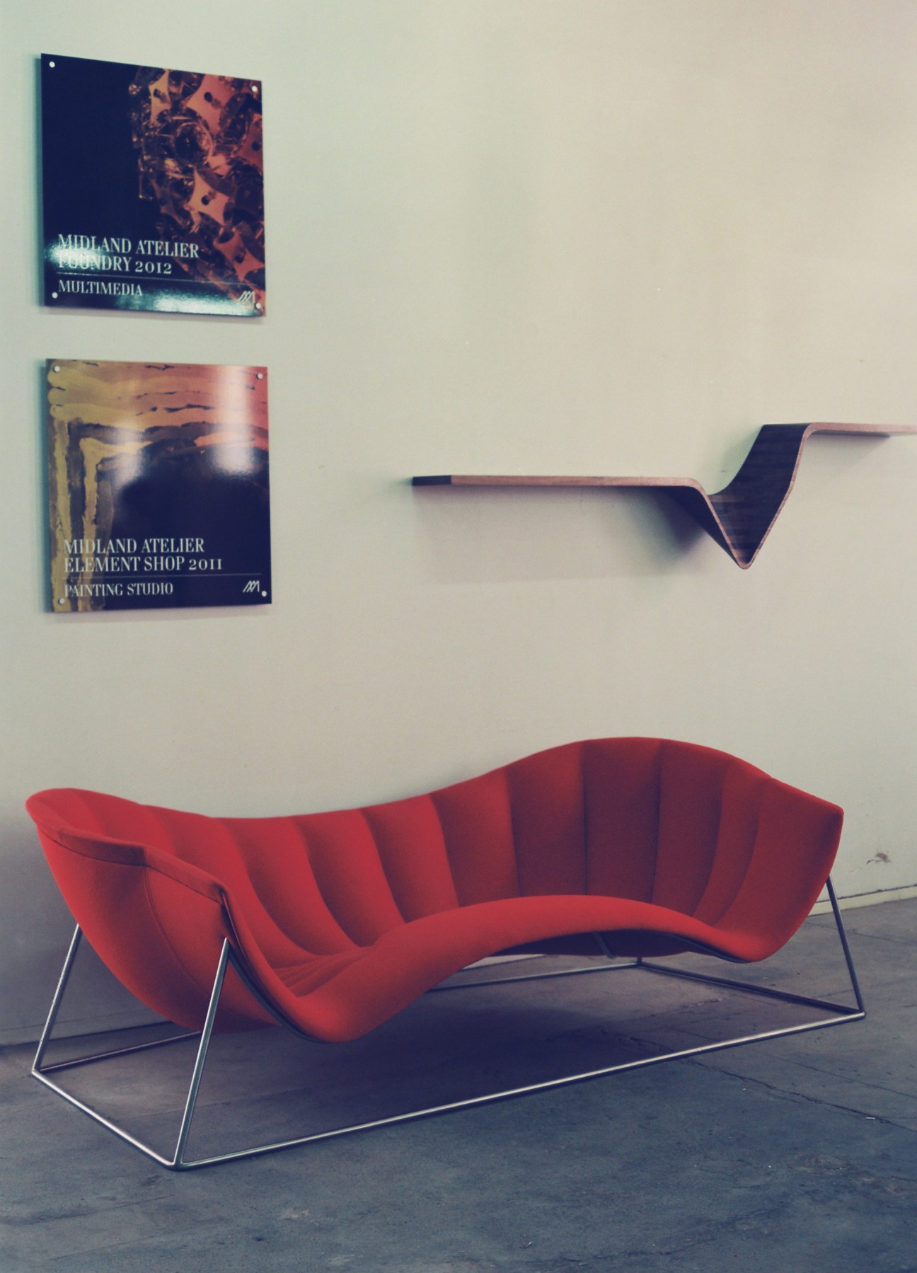 Red curved lounger chair with metal frame displayed in a gallery space with wall-mounted signage and sculptural shelf.