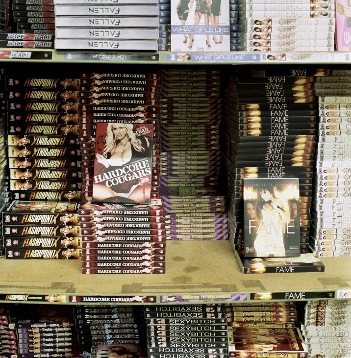 DVD cases of adult films stacked on shelves, including titles like 'Hardcore Cougars', 'Flashpoint', and 'Fame'.