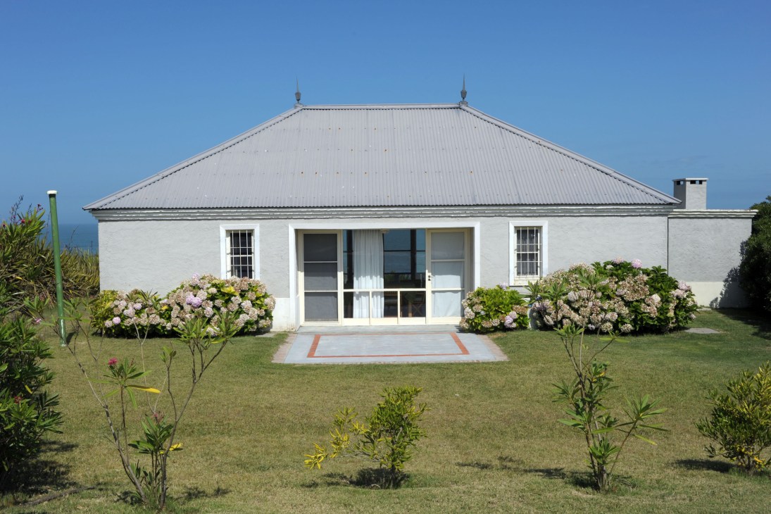 White single-story house with metal roof and large windows facing a manicured lawn with flowering shrubs.