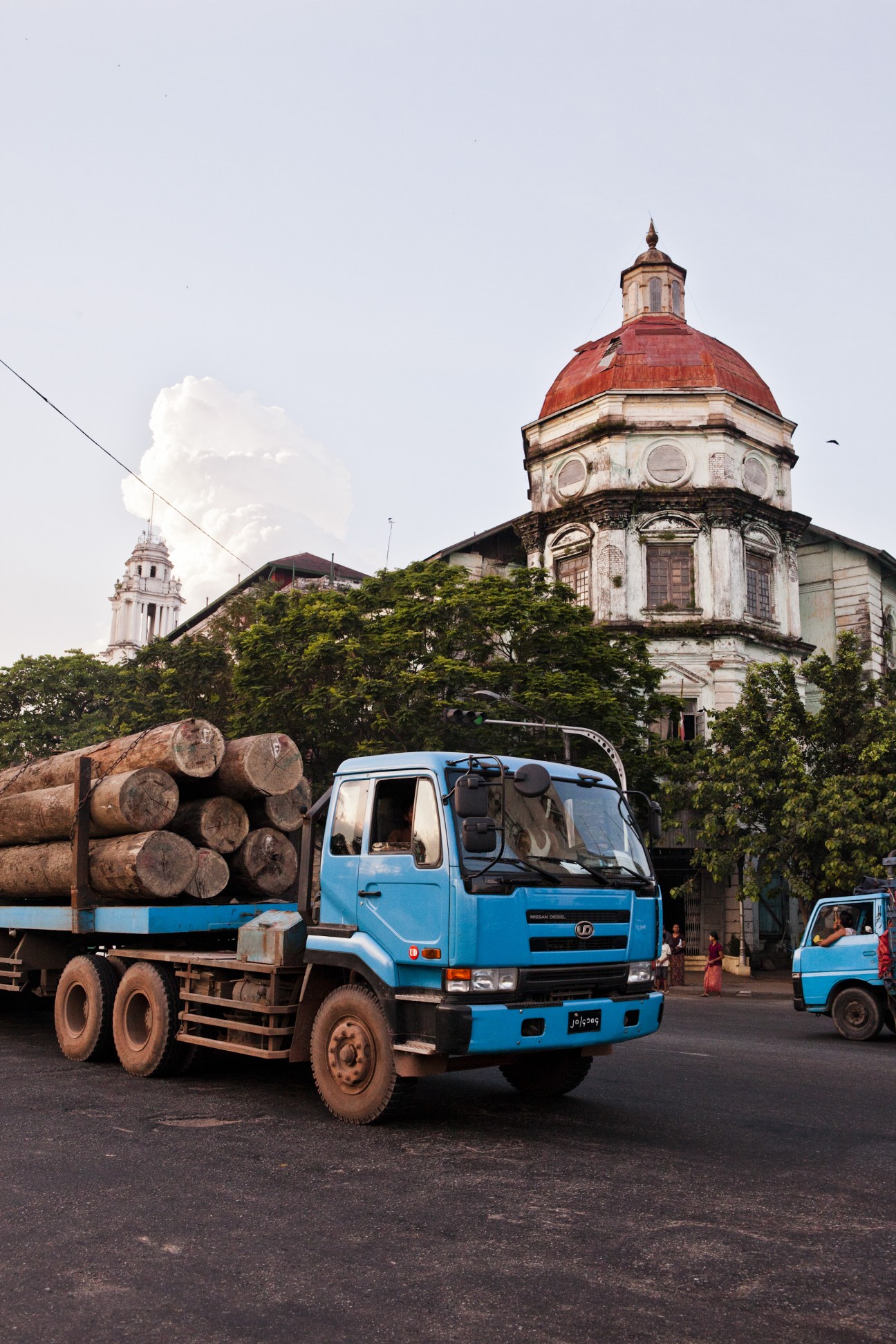 Blue logging truck loaded with timber passes colonial buildings with red dome on The Strand in Rangoon.