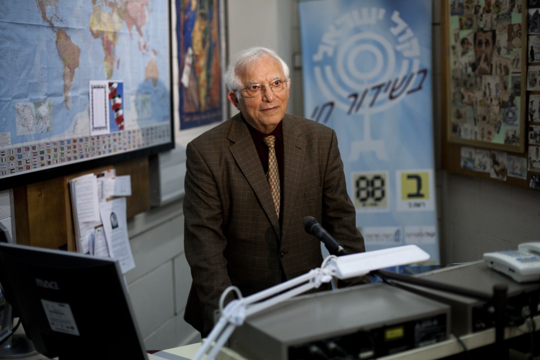 Menashe Amir, elderly man in brown jacket and glasses, speaking at microphone in radio studio with maps and documents visible behind him.