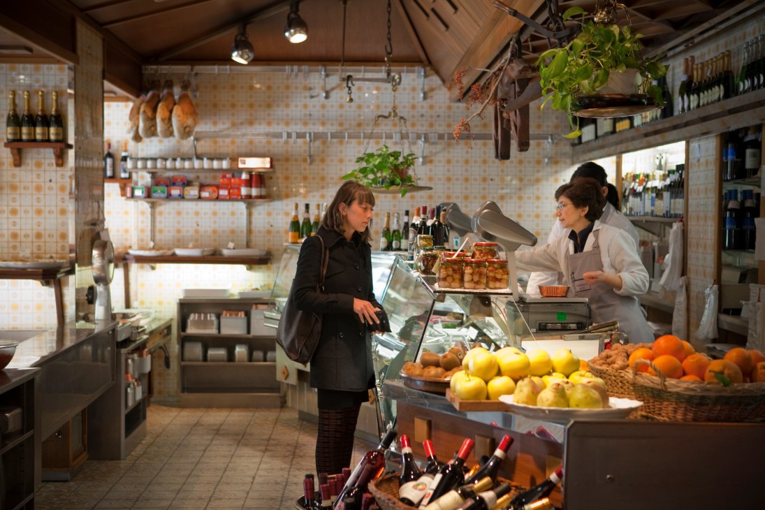 Interior of Baudracco deli in Turin showing a customer and shopkeeper near a display counter with fresh produce and wine bottles.