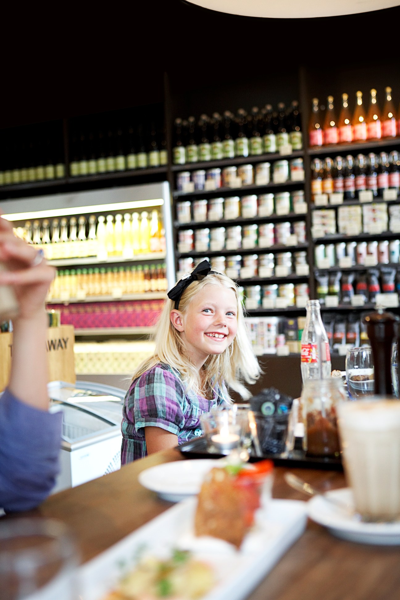 A smiling young girl with a black bow in her hair sits at a counter at Meyers Deli, with shelves of products visible behind her.