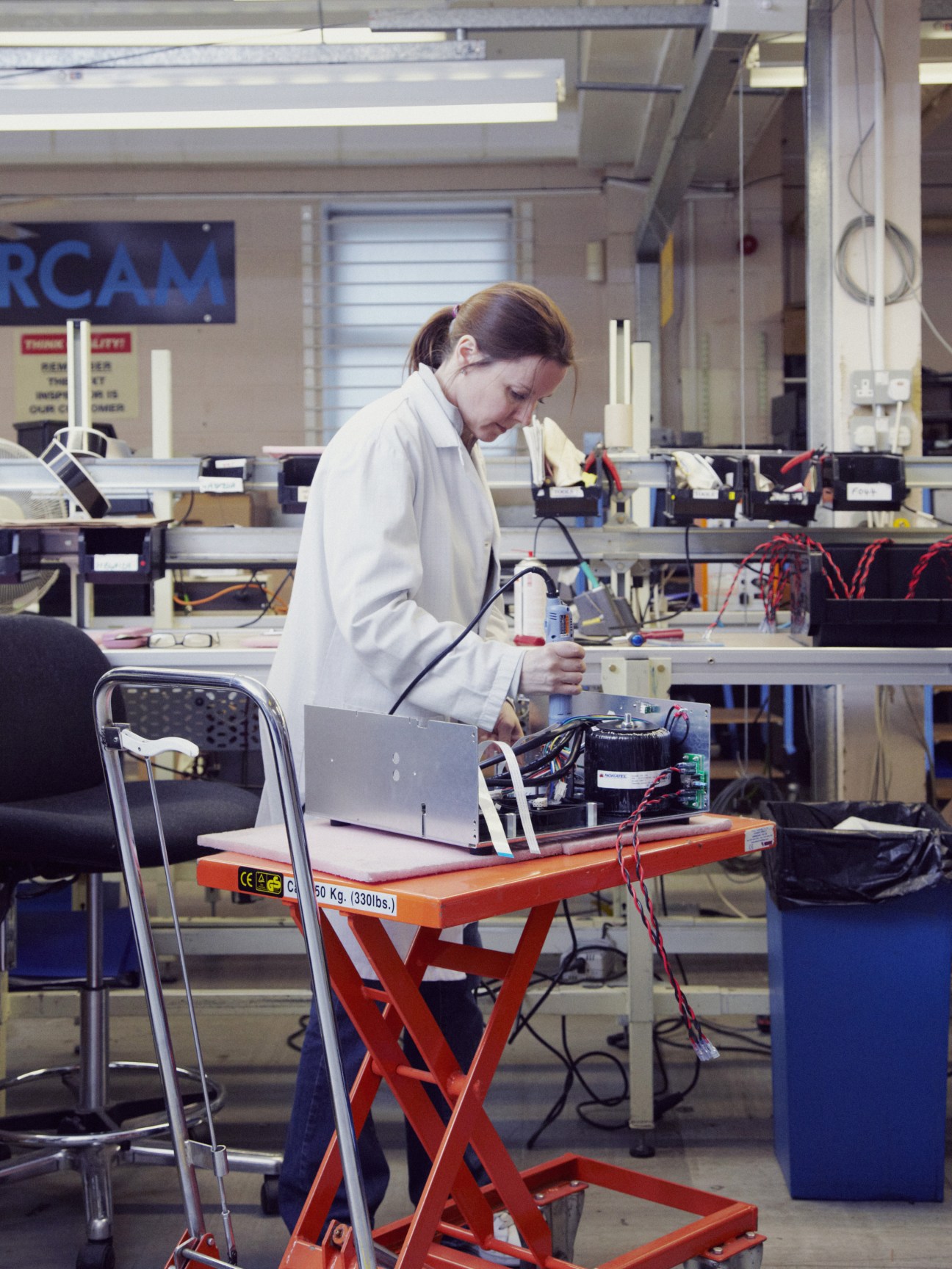 Technician in white coat tests audio receiver circuitry on red work platform in Arcam factory.