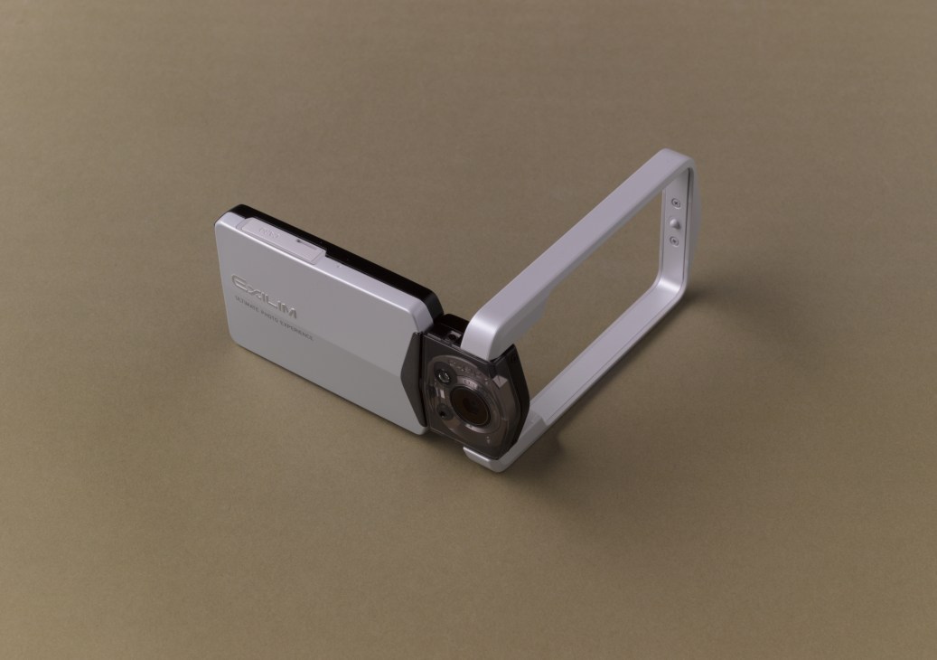 Casio Tryx EX-TR100 camera with rotating frame and extended arm on beige background.