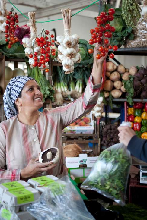 A vendor at a market stall reaches toward hanging bunches of garlic and red currants strung on green twine.