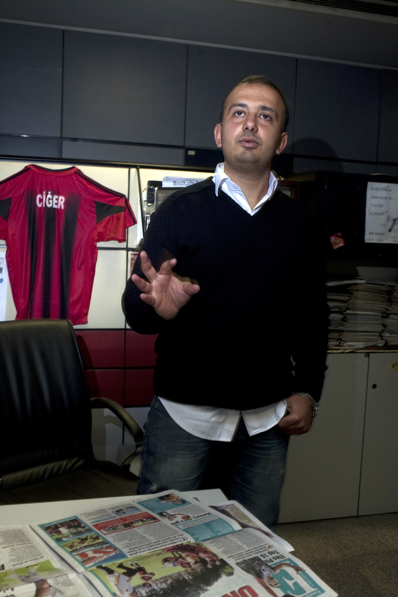 Kaan Bora, editor at Fanatik sports daily, speaking in an office with a red football jersey displayed behind him.