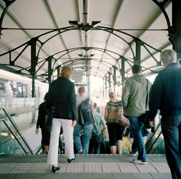Commuters walking through a covered bridge with green steel framework at Hilversum Noord station.