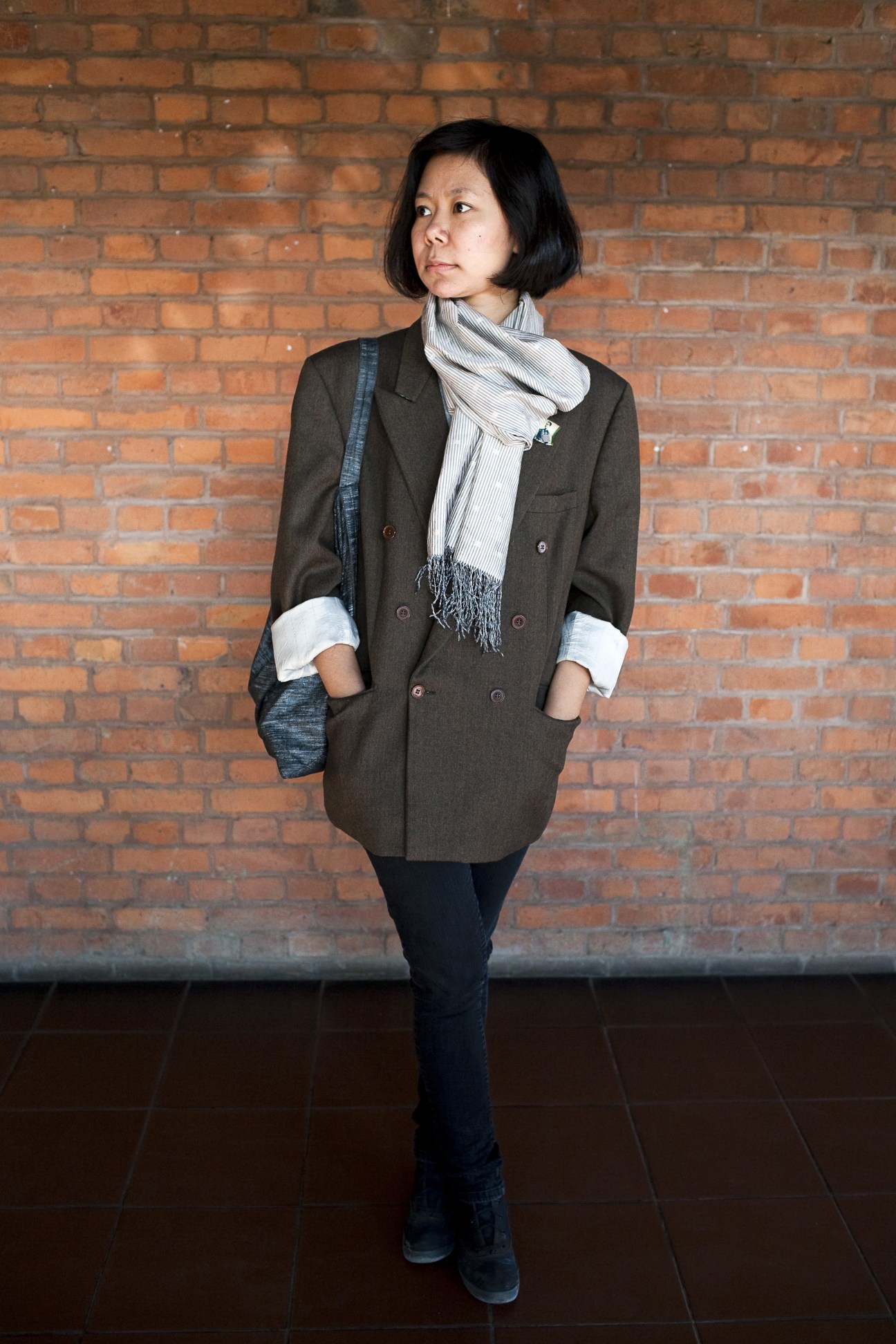 Designer Eiei Kyaw stands against a brick wall wearing a brown blazer and striped scarf.