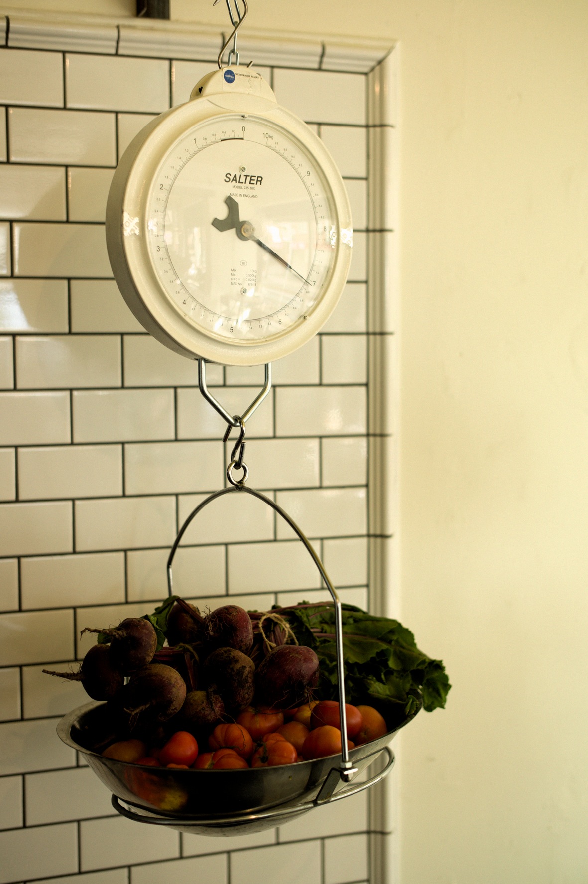 A vintage Salter hanging scale with fresh produce in the basket against subway tile wall.