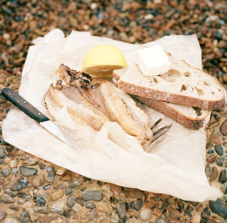 Smoked kipper fillets with lemon, brown bread, butter and knife on pebbled beach.