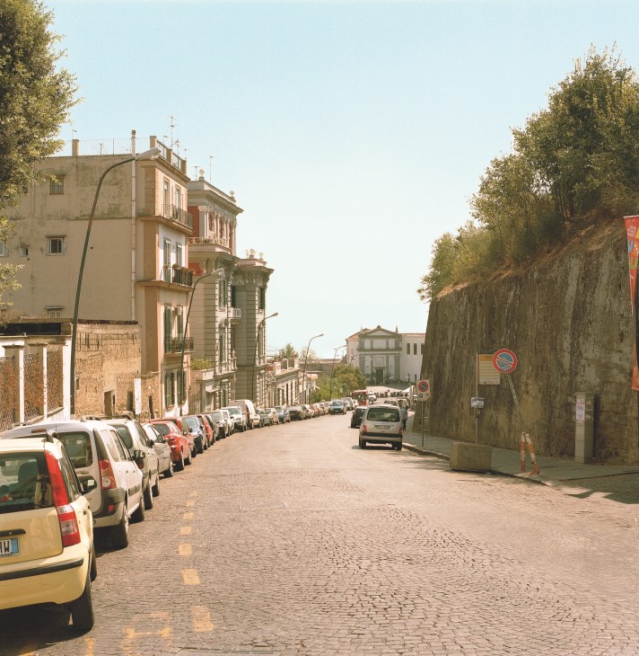 Tree-lined street in Naples with parked cars and early 20th-century Liberty-style buildings.