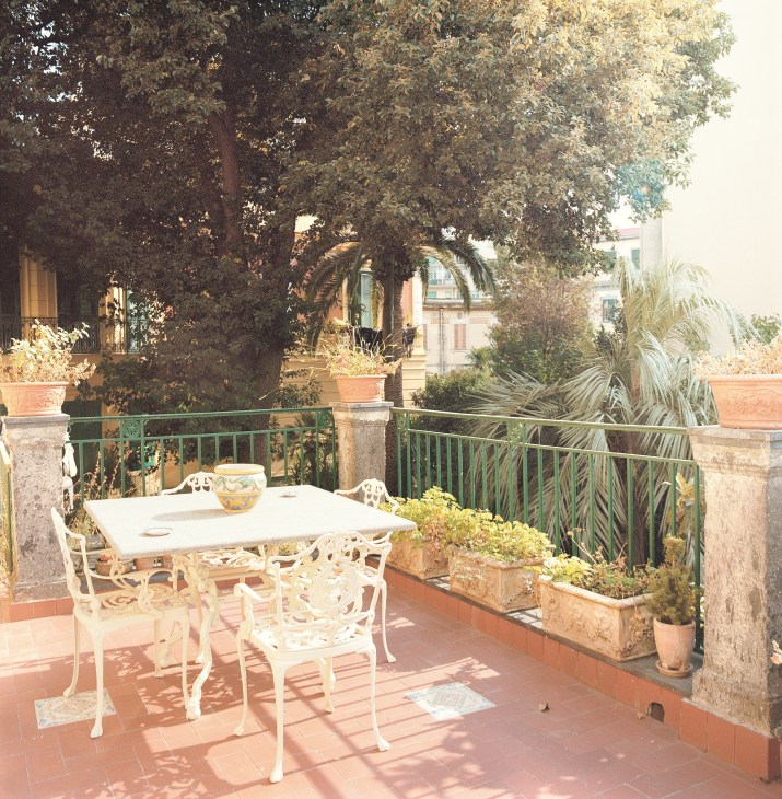 A sunny terrace with white metal furniture, potted plants, and green railing overlooking Naples.