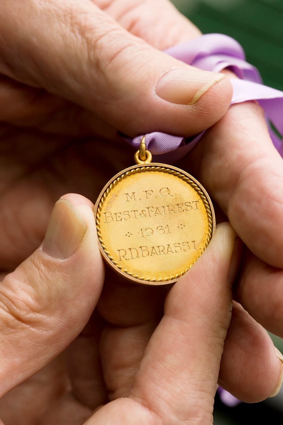 Hand holding a gold medal engraved with 'M.F.C. BEST&FAIREST 1961 R.D.BARASSI' on a purple ribbon.