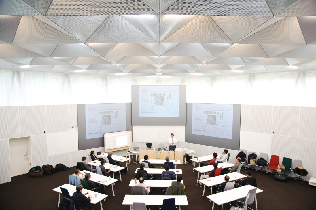 Training room with angular white ceiling, projector screens, and participants seated at tables with colorful chairs.
