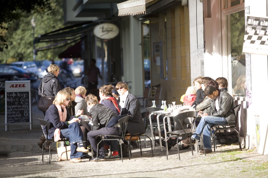 People seated at outdoor café tables on a sunny Neukölln street with pre-war buildings.