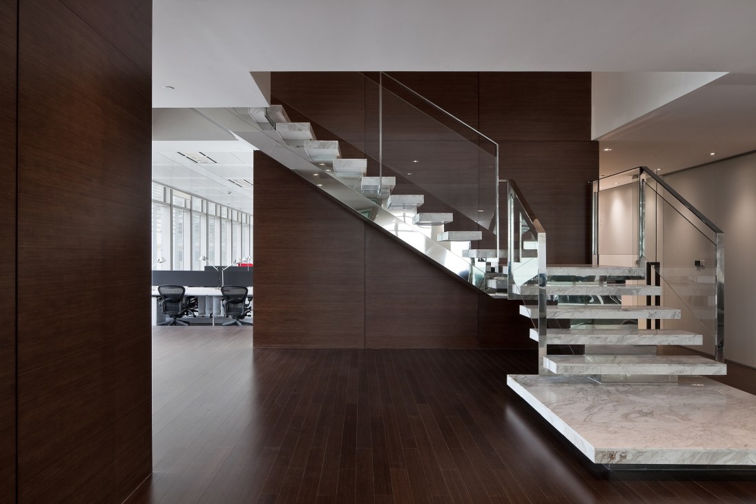 Modern office staircase with dark wood paneling, marble steps, and glass railings overlooking workspace.