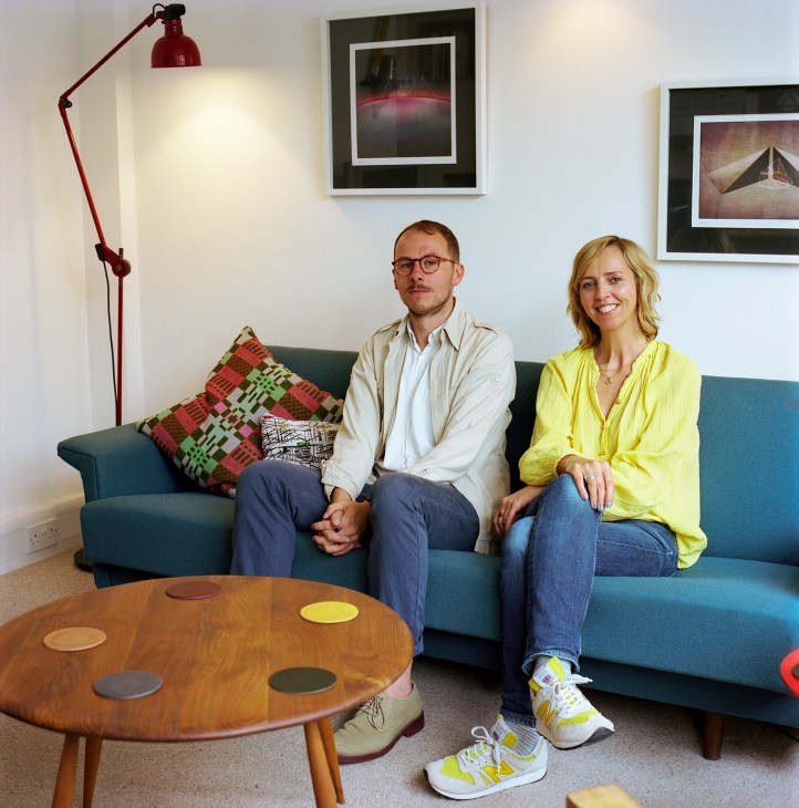 Two people sit on a teal sofa in a mid-century modern office interior with a red floor lamp and framed artwork.