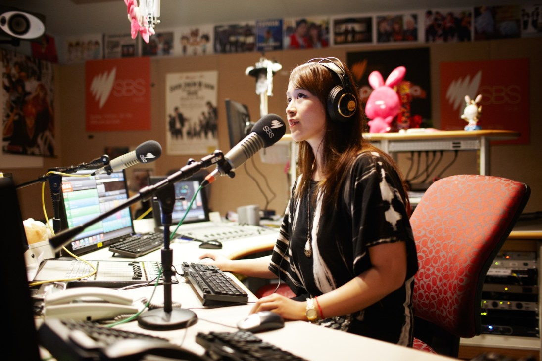 Jamaica dela Cruz hosting PopAsia on SBS Radio, speaking into a microphone in a studio decorated with posters and pink decorative items.