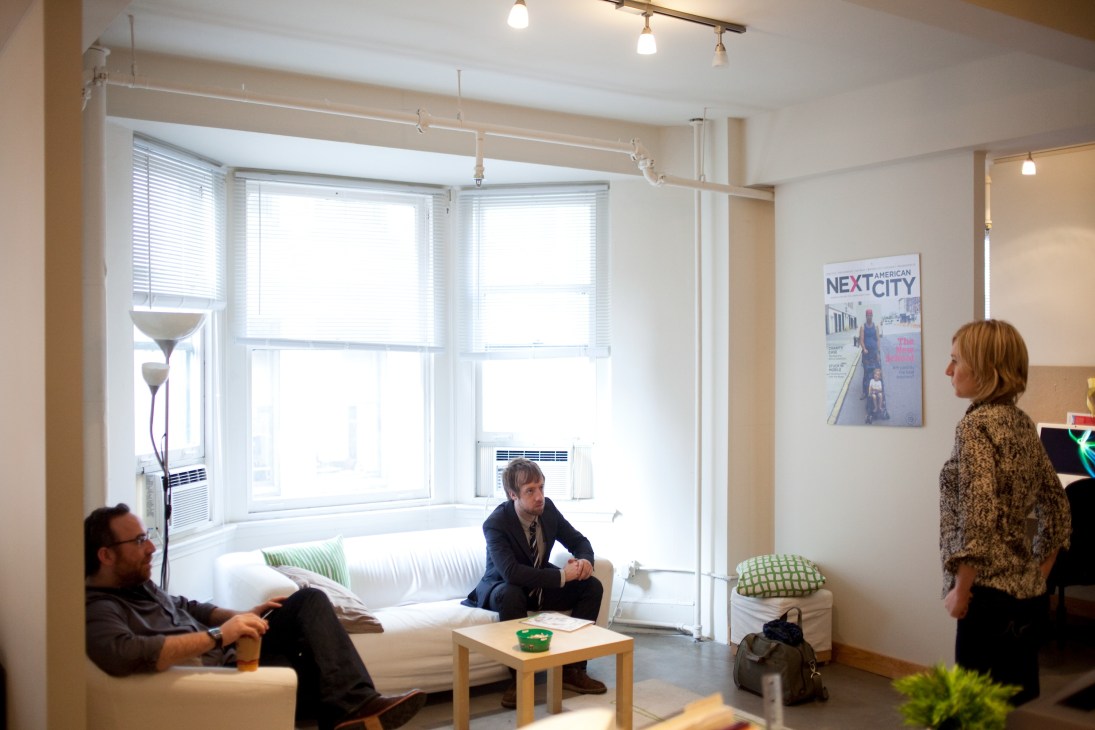 Three people in a modern office space with white furniture, large windows, and a Next American City magazine cover on the wall.